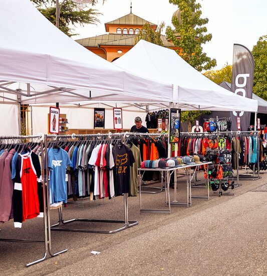 The white promotional tents serve as a sales tent at an event. Various t-shirts hang under the folding gazebo.