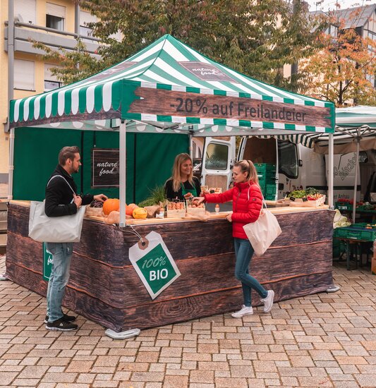 The market tent is fully printed and is used at the weekly market and farmers' market. 2 customers look at the products.