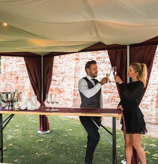 Under the elegant folding gazebo with a sky, 2 people stand with a glass of prosecco in their hands. 