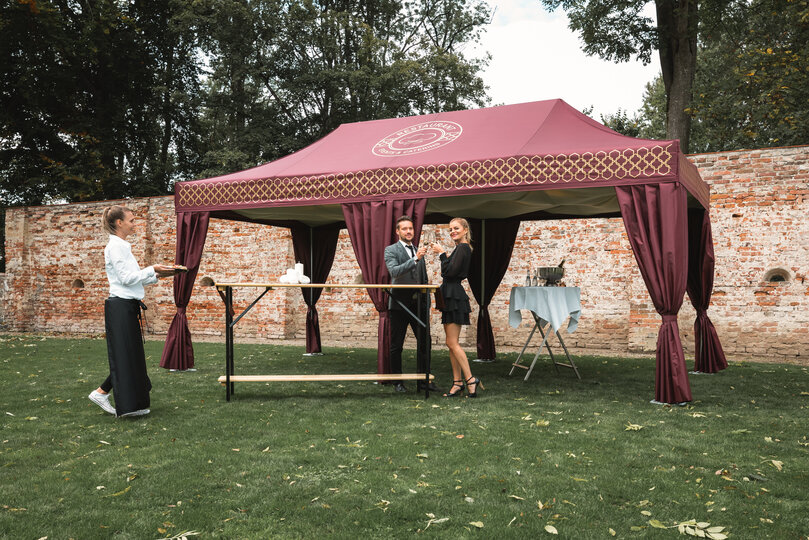 The bordeaux-coloured catering tent stands in a meadow. A couple is standing in front of it holding a glass of prosecco. The waitress brings appetisers on a tray.