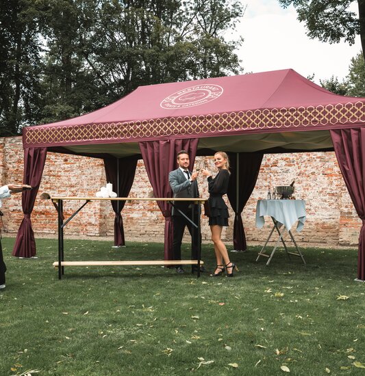 The bordeaux-coloured catering tent stands in a meadow. A couple is standing in front of it holding a glass of prosecco. The waitress brings appetisers on a tray.