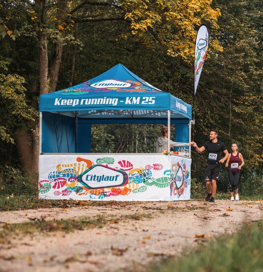 Blue 3x3m event gazebo with custom Citylauf print, with custom flag, closed side wall and half-height side wall with counter used as a refreshment stand at a city race